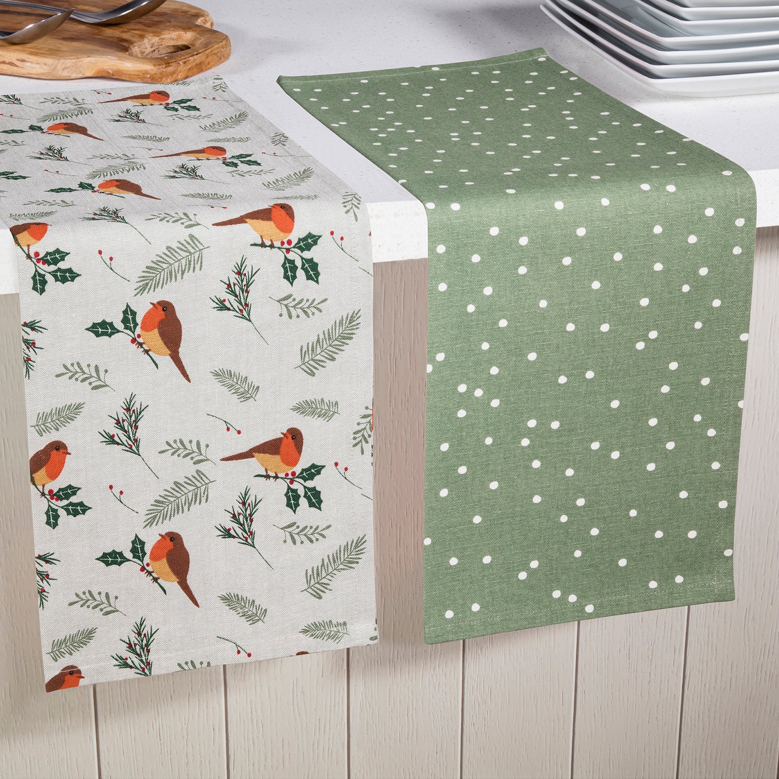 Two table runners draped over a white kitchen counter. One runner has a light gray background with a pattern of orange birds, green holly leaves, and pine branches.