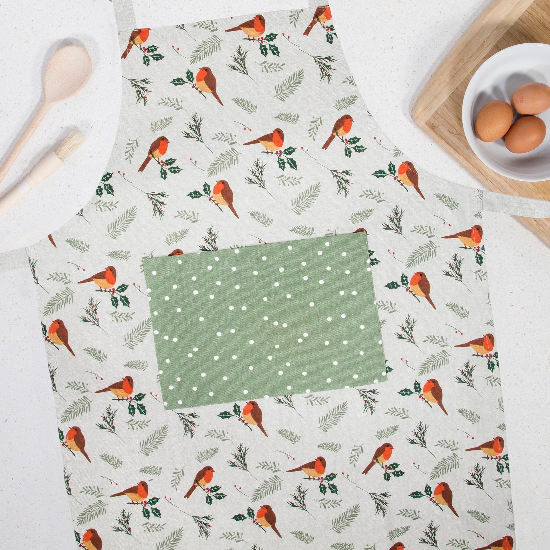 A flat-lay view of a kitchen apron with a white background, printed with orange birds and green leaves, featuring a solid sage green pocket with white polka dots. The apron is styled on a white countertop with a wooden spoon, a wooden cutting board, a small white bowl of eggs, and a striped kitchen towel.