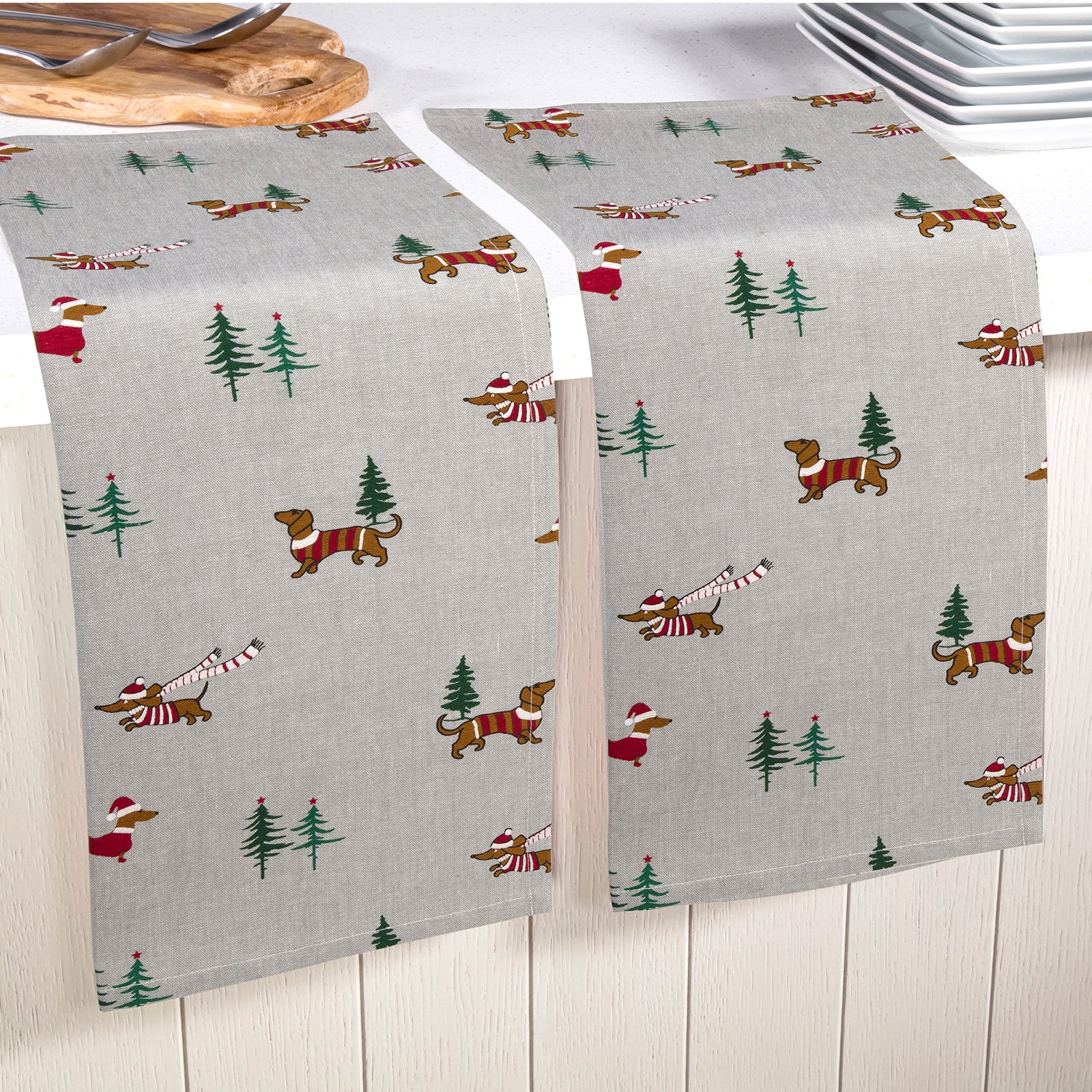 Two light gray table runners with a dachshund and Christmas tree pattern, draped over a white table in a kitchen setting.