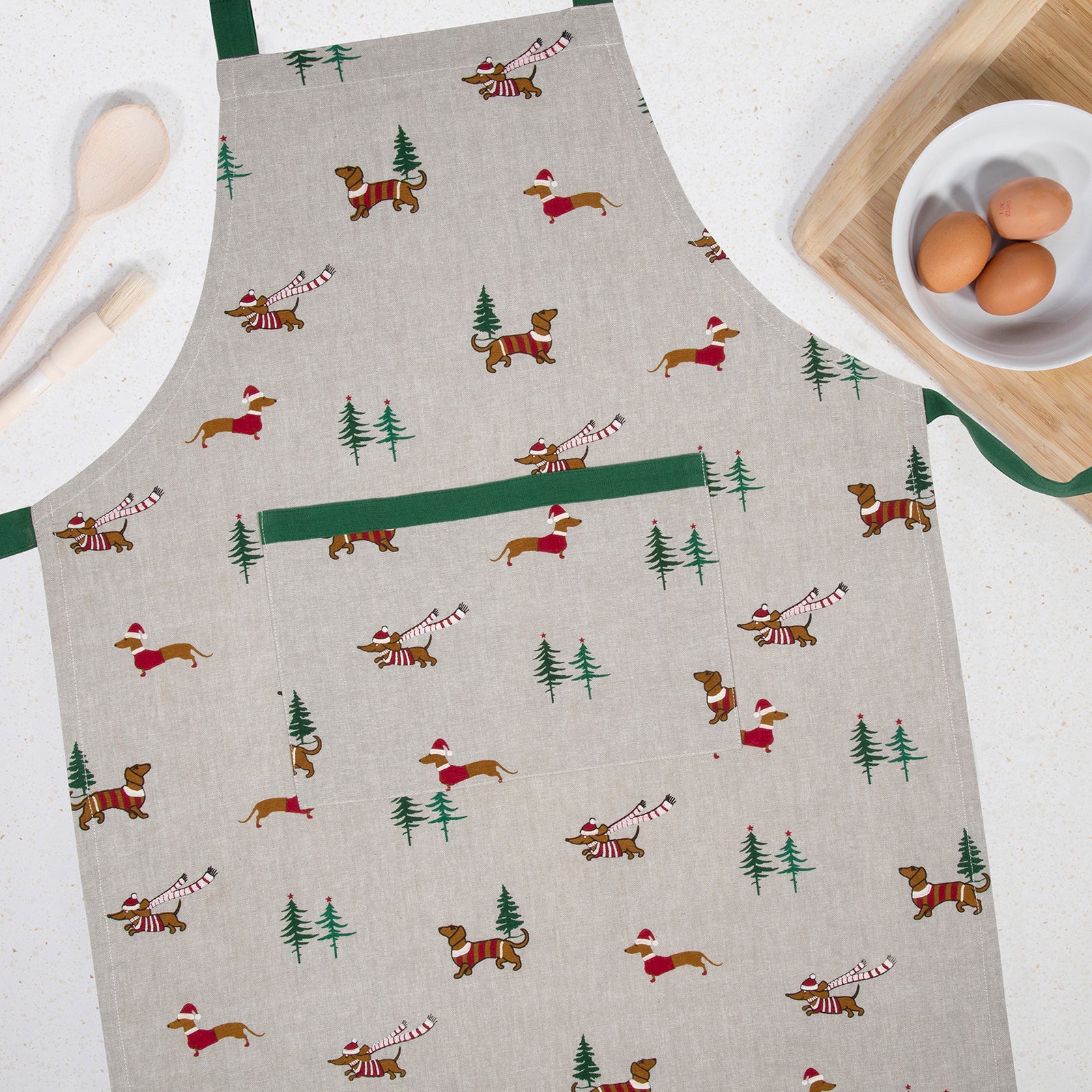 A light beige linen apron with a green neck strap and waist loop, featuring a festive print of dachshunds in red Santa hats, Christmas trees, and candy canes. The apron is styled on a white countertop with kitchen props including wooden spoons, a cutting board, and a bowl of eggs.