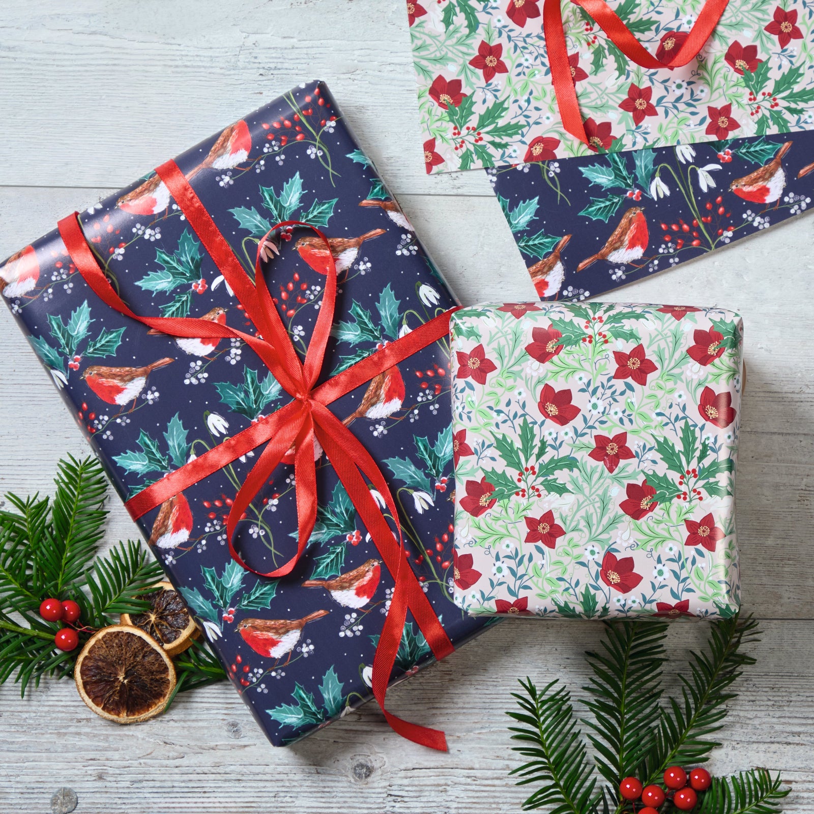 Three wrapped Christmas gift boxes arranged on a light gray wood surface with evergreen branches, red berries, and dried orange slices. The wrapping paper features festive holiday prints including birds, holly, berries, and floral motifs, with one large box tied in the center by a red ribbon.