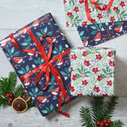 Three wrapped Christmas gift boxes arranged on a light gray wood surface with evergreen branches, red berries, and dried orange slices. The wrapping paper features festive holiday prints including birds, holly, berries, and floral motifs, with one large box tied in the center by a red ribbon.